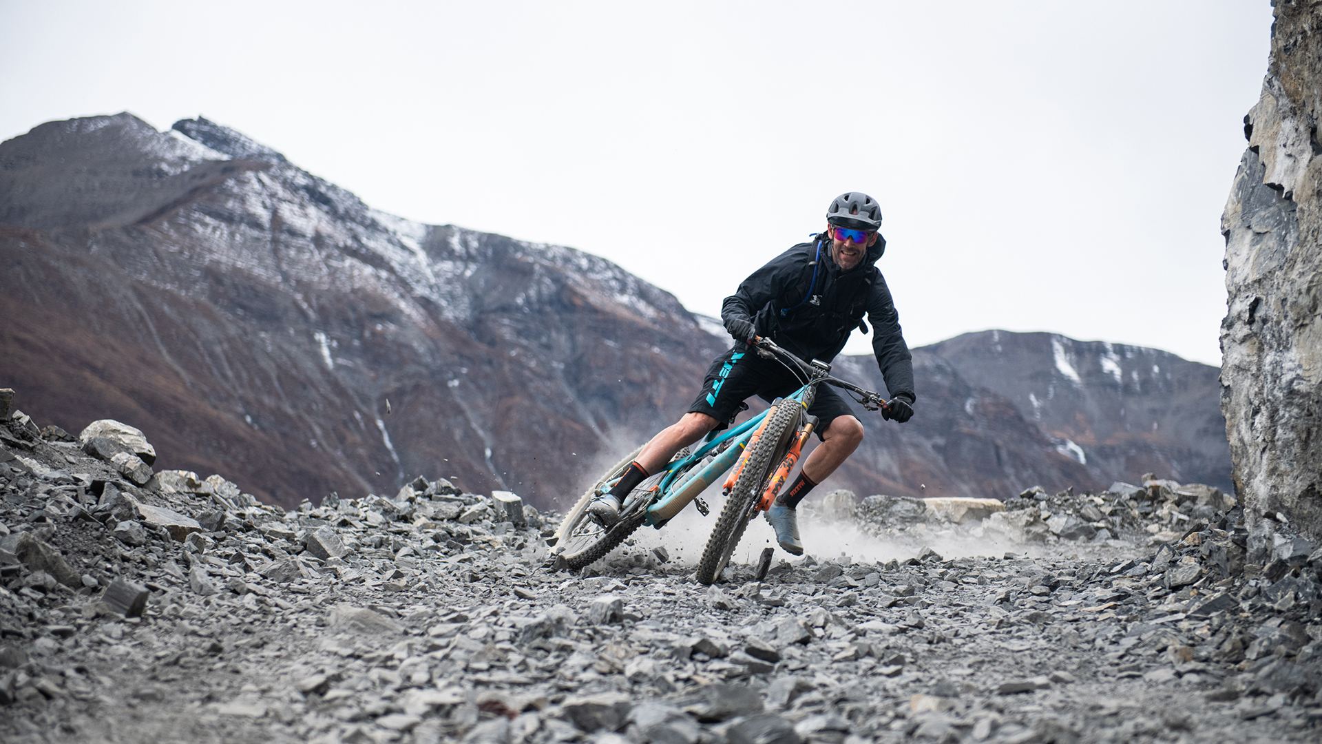 A rider leans into a corner on grey scree with mountains behind