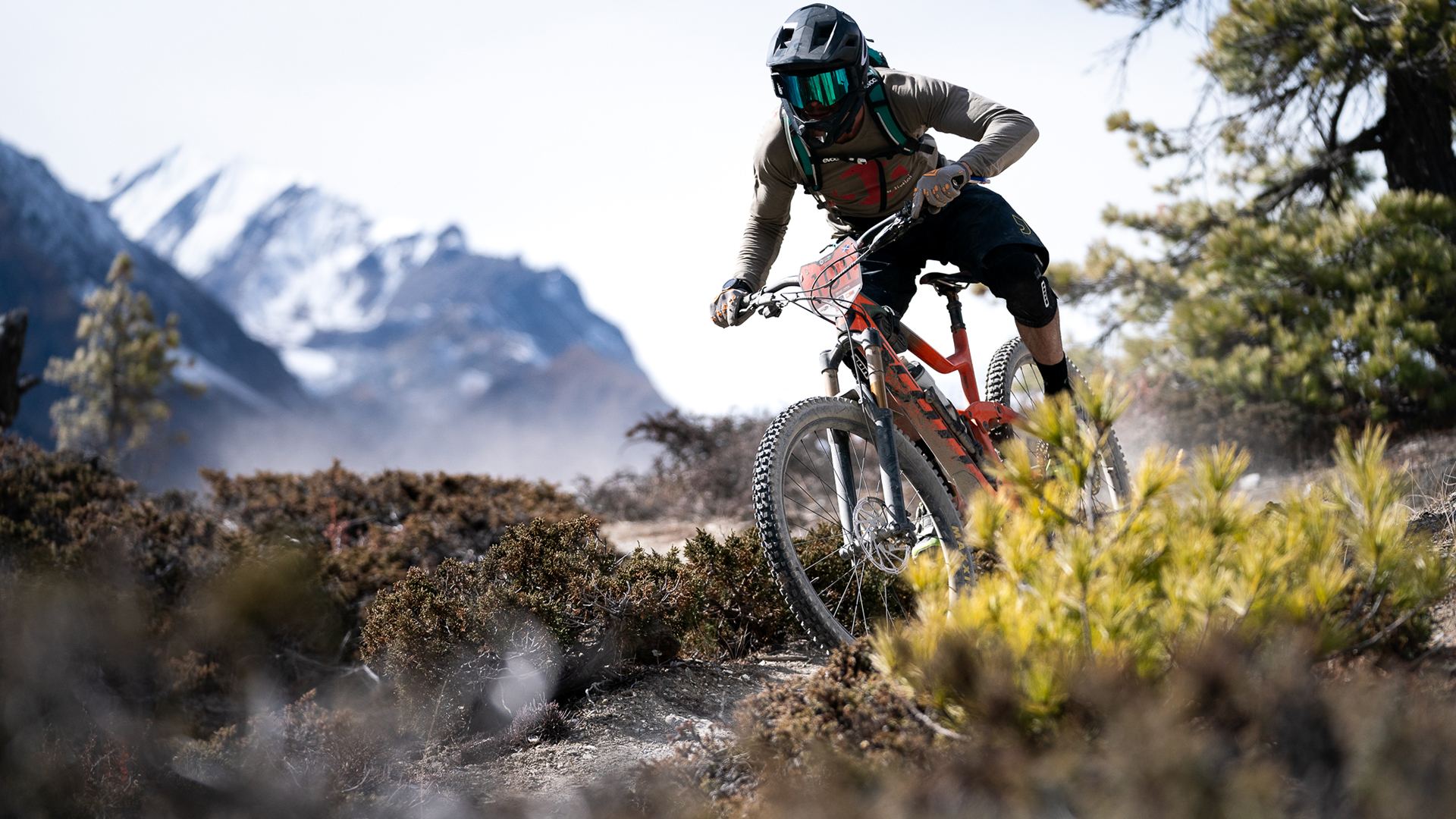 A rider carves a dusty Himalayan trail with snow peaks behind